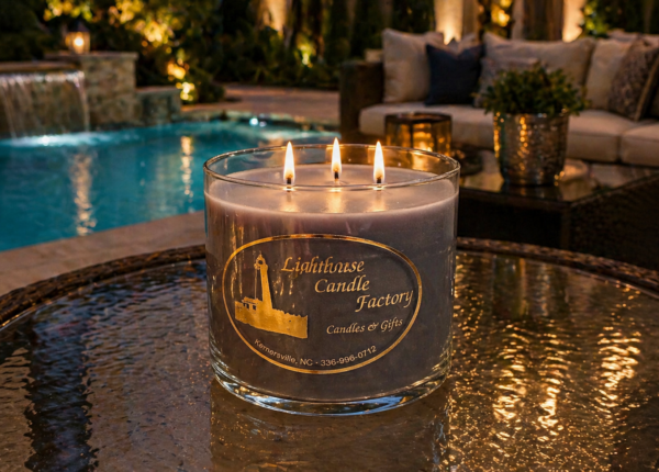 Luxury three-wick candle from The Lighthouse Candle Factory on glass table by swimming pool with outdoor patio furniture at night