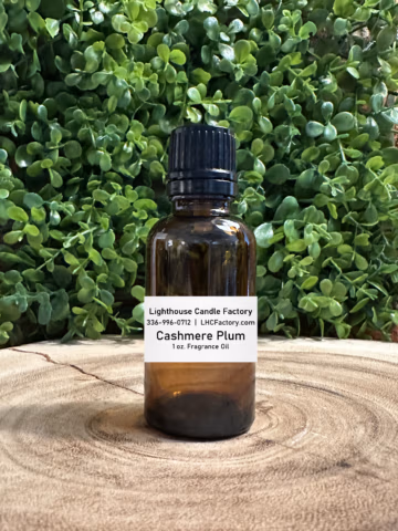 1 oz. Cashmere Plum fragrance oil by The Lighthouse Candle Factory in an amber glass bottle with a black, tamper-evident dropper cap, set against a greenery background.