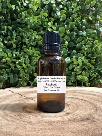 1 oz. Patchouli Odor Be Gone (OBG) fragrance oil by The Lighthouse Candle Factory in an amber glass bottle with a black, tamper-evident dropper cap, set against a greenery background.