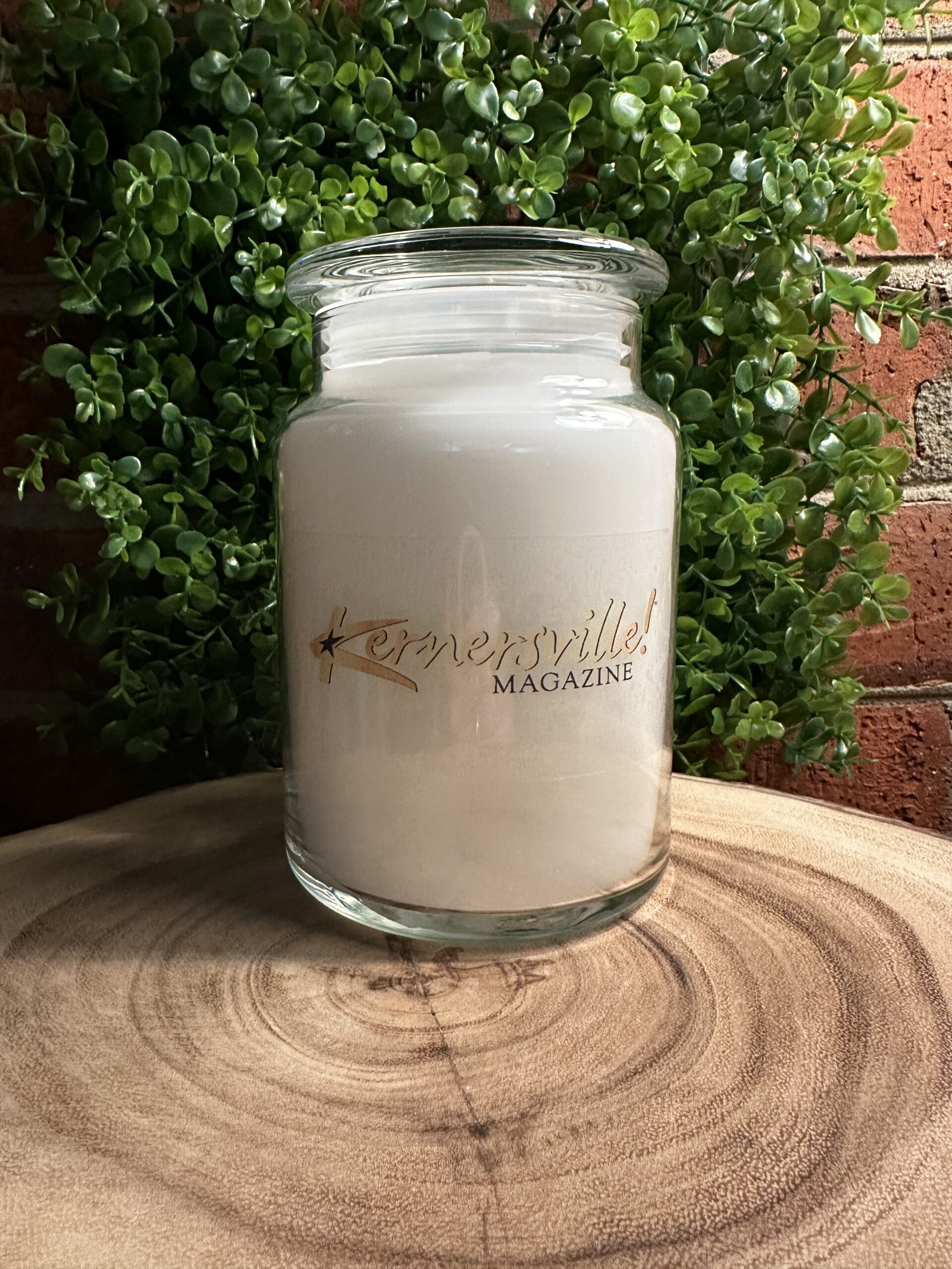 One of The Lighthouse Candle Factory's Custom Corporate Candle Gifts inside a clear glass jar with white wax featuring the Kernersville Magazine logo.