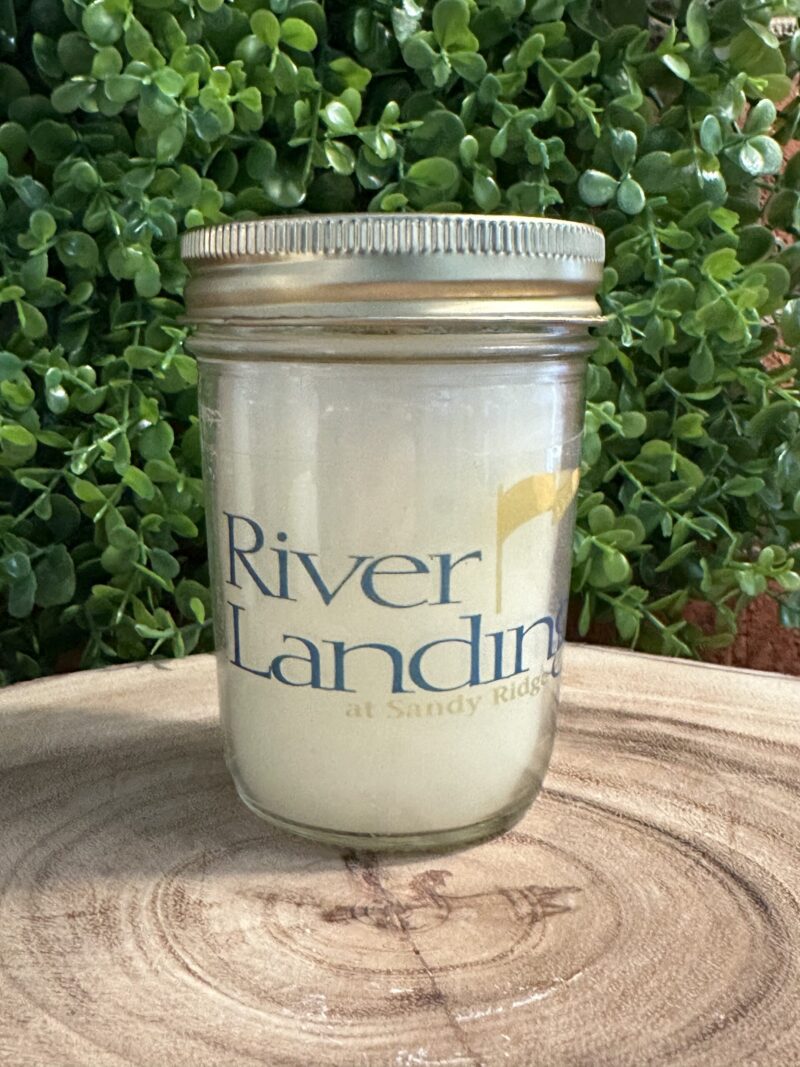 One of The Lighthouse Candle Factory's Custom Corporate Candle Gifts inside a clear glass jar with white wax featuring the River Landing Retirement Community logo.