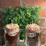 Two realistic handmade Root Beer Float candles in authentic A&W glass mugs with whipped wax foam tops, styled as a custom engagement gift in front of a brick wall and greenery.