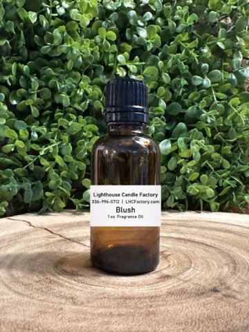 1 oz. Blush fragrance oil by The Lighthouse Candle Factory in an amber glass bottle with a black, tamper-evident dropper cap, set against a greenery background.
