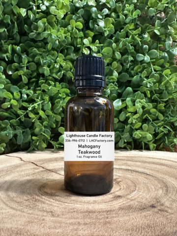 1 oz. Mahogany Teakwood fragrance oil by The Lighthouse Candle Factory in an amber glass bottle with a black, tamper-evident dropper cap, set against a greenery background.