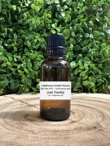 1 oz. Just Vanilla fragrance oil by The Lighthouse Candle Factory in an amber glass bottle with a black, tamper-evident dropper cap, set against a greenery background.