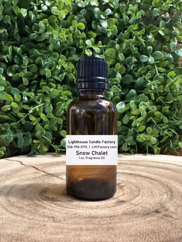 1 oz. Snow Chalet fragrance oil by The Lighthouse Candle Factory in an amber glass bottle with a black, tamper-evident dropper cap, set against a greenery background.