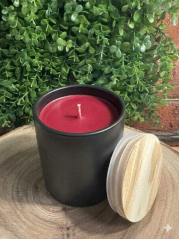 Apple Bourbon Candle unlit in a black ceramic vessel with red wax and a wood-grain lid leaning beside it.