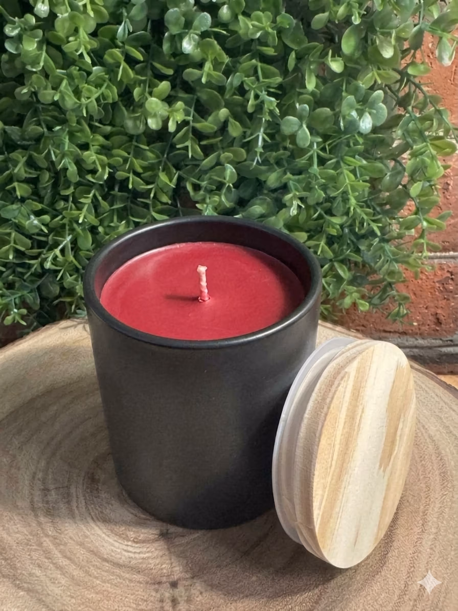 apple_bourbon_black_12oz_modern_ceramic_tumbler Apple Bourbon Candle unlit in a black ceramic vessel with red wax and a wood-grain lid leaning beside it.