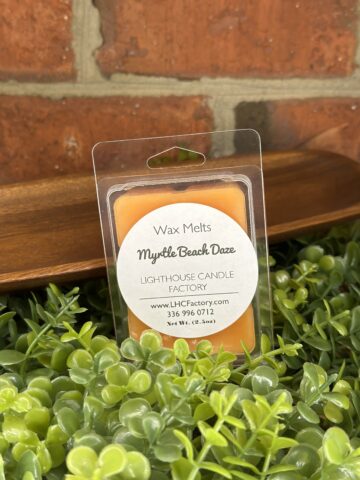 A 2.5oz clamshell package of orange Myrtle Beach Daze wax melts by The Lighthouse Candle Factory, resting on a bed of green faux foliage.