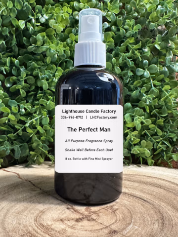 An 8 oz. dark amber spray bottle of "The Perfect Man" All Purpose Fragrance Spray by The Lighthouse Candle Factory, centered against a dense background of green leafy foliage on a rustic wooden slice.