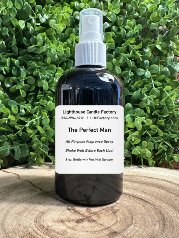 An 8 oz. dark amber spray bottle of "The Perfect Man" All Purpose Fragrance Spray by The Lighthouse Candle Factory, centered against a dense background of green leafy foliage on a rustic wooden slice.