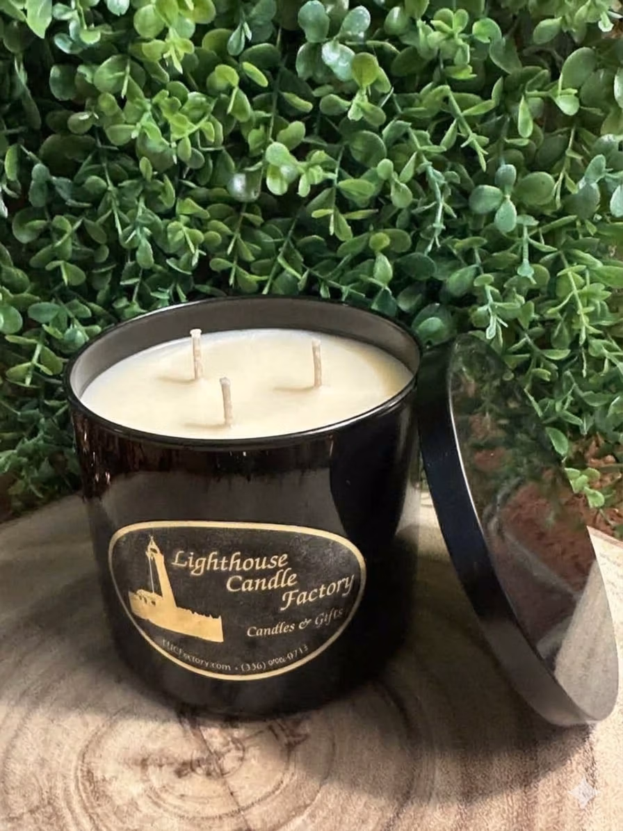 amazing_grace_3_wick_black_top Amazing Grace floral musk candle in a black vessel with three unlit wicks and a lid leaning beside it, against green foliage.