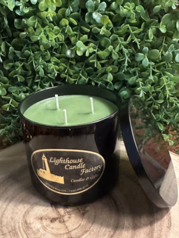 Bayberry scented candle in a black vessel with three unlit wicks, green wax and a lid leaning beside it against leafy greenery.