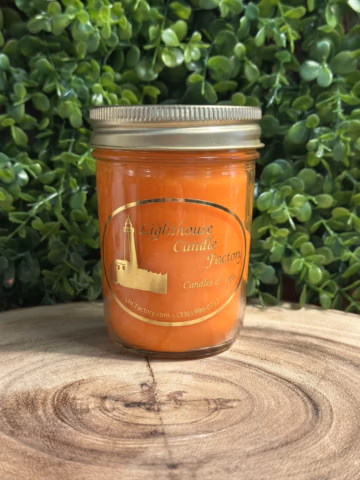 Front view of pumpkin pie scented candle with a gold lid and Pumpkin Orange wax, on a wood slice against green foliage.